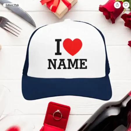 A white and navy trucker cap with the text "I ♥ NAME" in black and red, shown on a romantic-themed table with roses and a ring box.